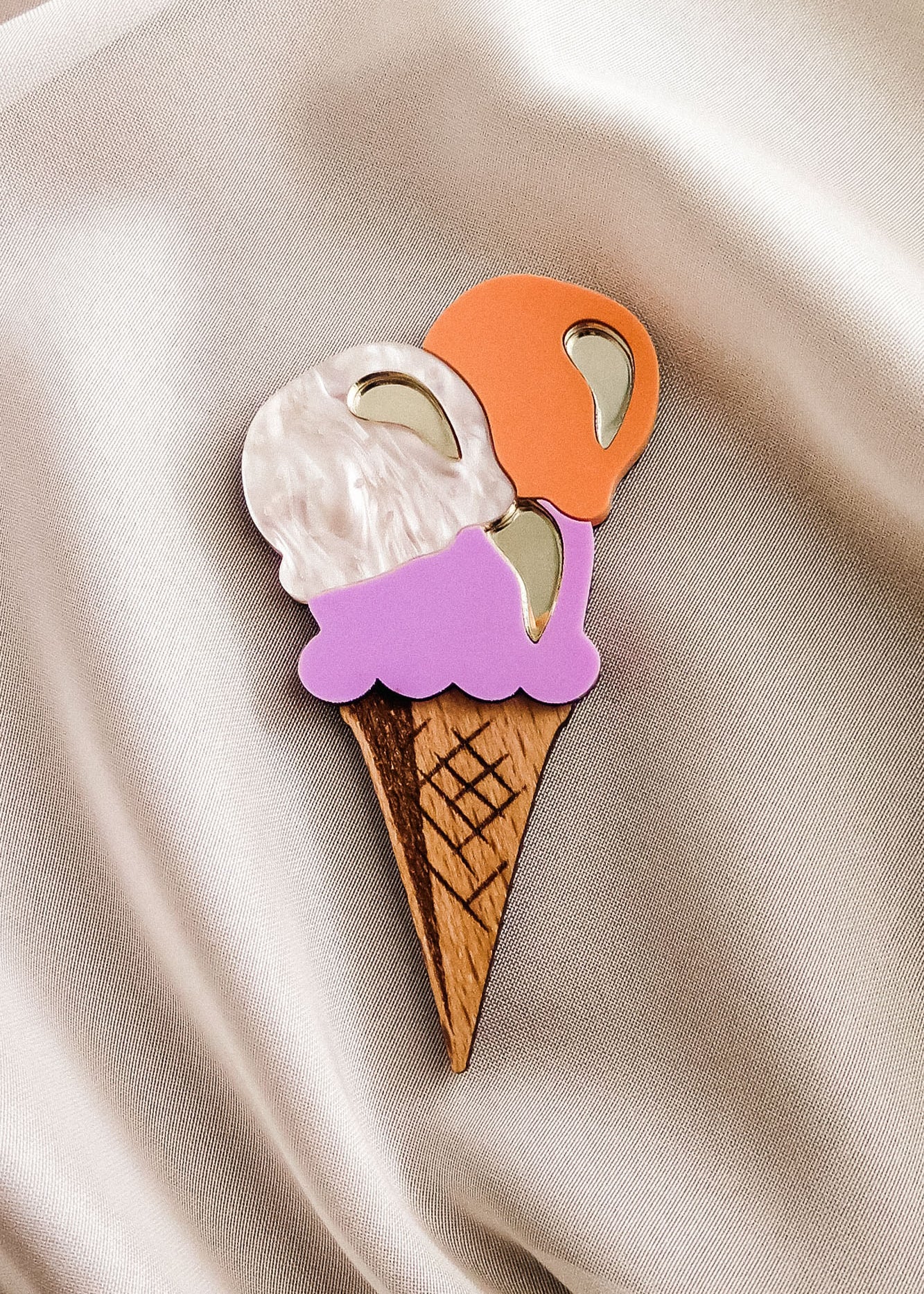 A handcrafted wooden ice cream brooch with white, orange, and purple scoops and shiny metallic accents is placed on light beige satin fabric—perfect for elevating your summer look.