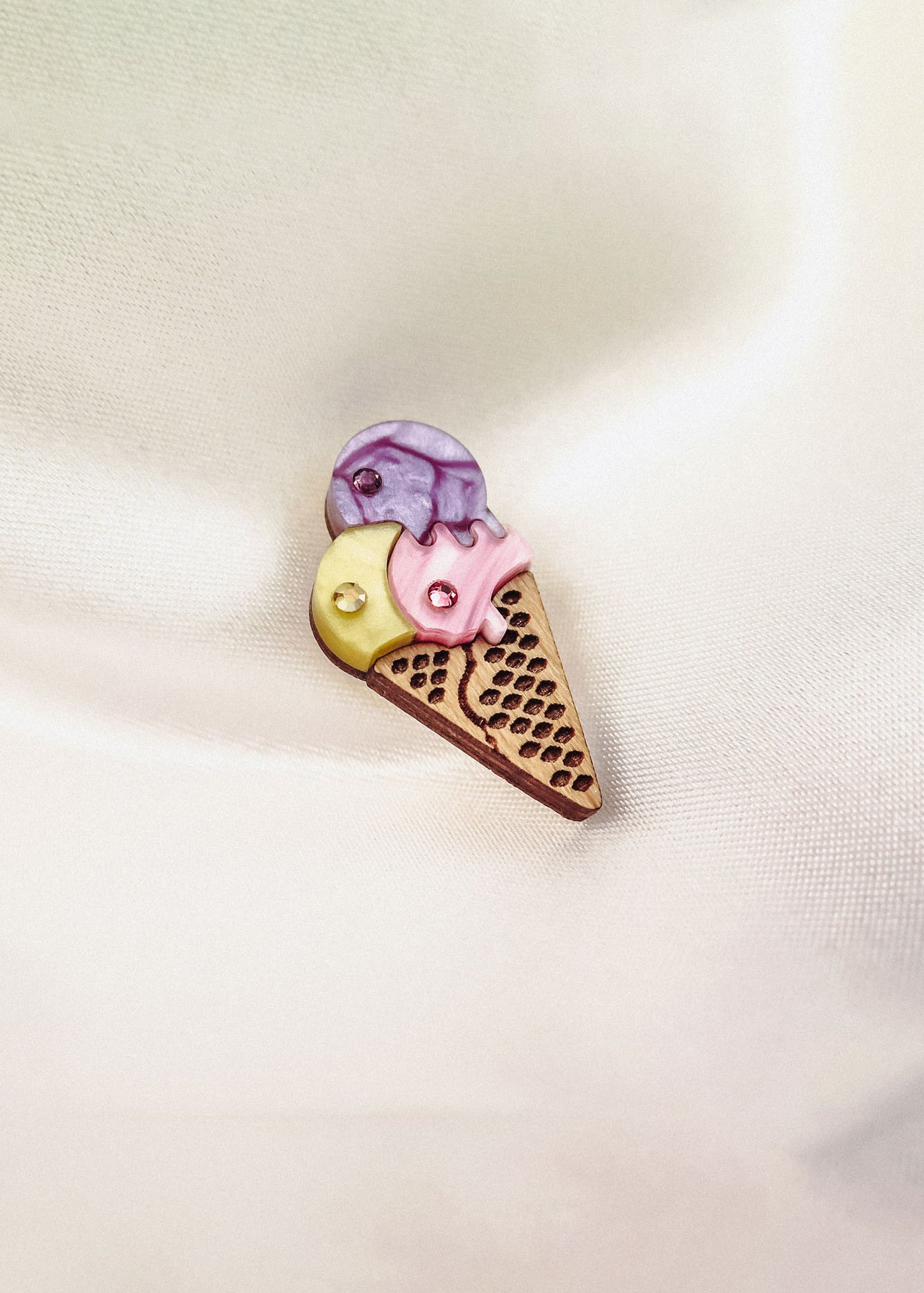 A handcrafted Ice Cream Pin shaped like a cone with three scoops—yellow, pink, and purple—features a playful design with small embedded rhinestones, resting on a soft, light-colored fabric background.