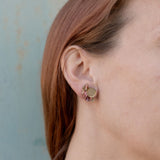 Close-up of a person with light skin and reddish-brown hair wearing a small, round, gold Minimalist Geometric Stud Earring featuring a wavy, pink and green patterned design on their left ear against a light blue background.