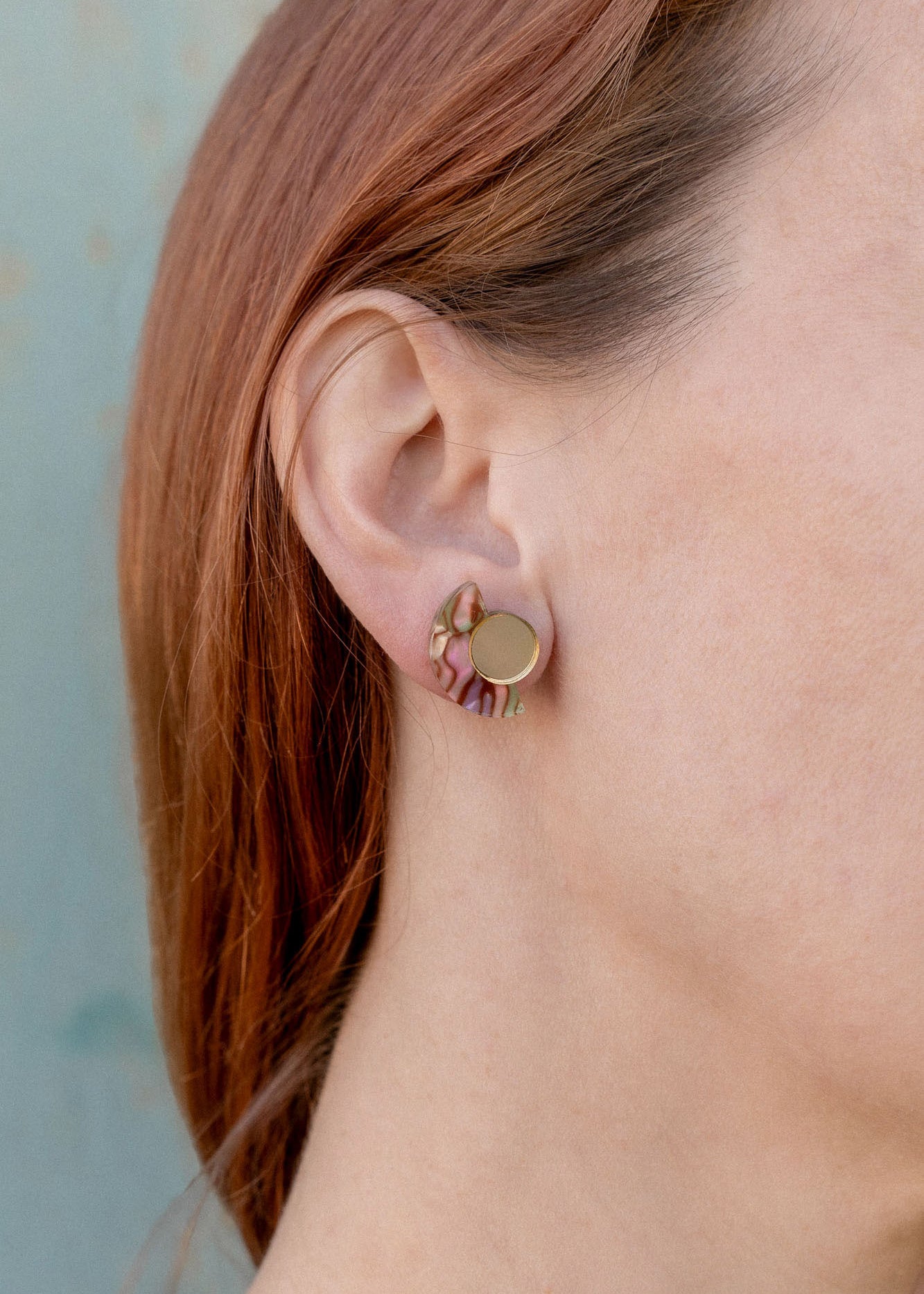 Close-up of a person with light skin and reddish-brown hair wearing a small, round, gold Minimalist Geometric Stud Earring featuring a wavy, pink and green patterned design on their left ear against a light blue background.