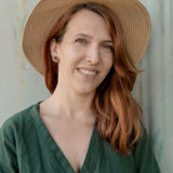A woman with long auburn hair wears minimalist geometric stud earrings, a wide-brimmed straw hat, and a green dress, smiling softly while standing in front of a neutral-colored wall.