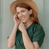 A woman with reddish-brown hair wears a green dress, a straw hat, and minimalist geometric stud earrings. She smiles gently at the camera while touching her face, standing in front of a light-colored background.