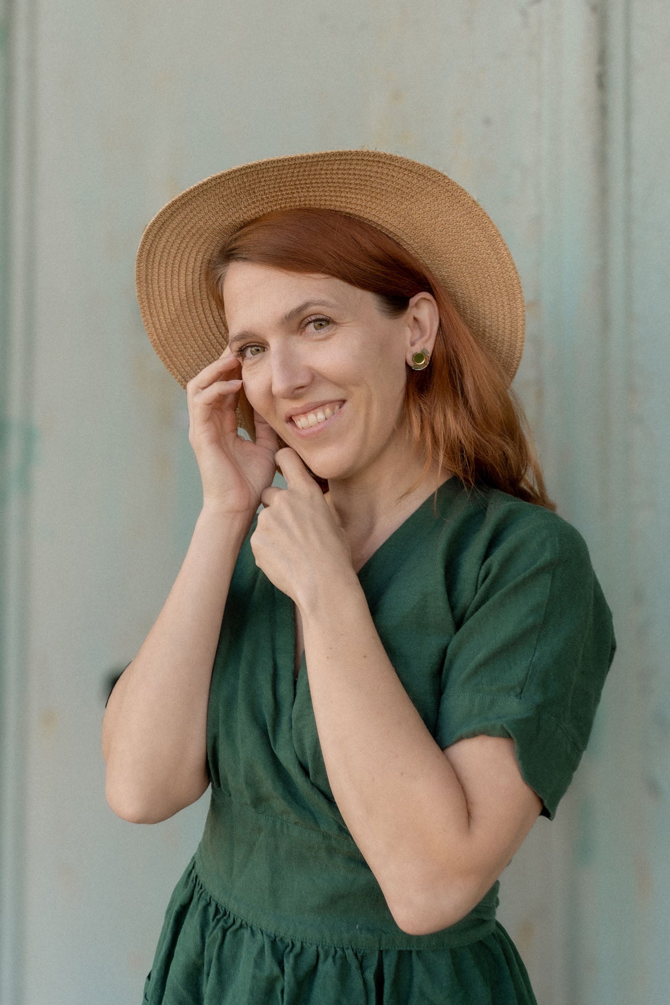 A woman with reddish-brown hair wears a green dress, a straw hat, and minimalist geometric stud earrings. She smiles gently at the camera while touching her face, standing in front of a light-colored background.