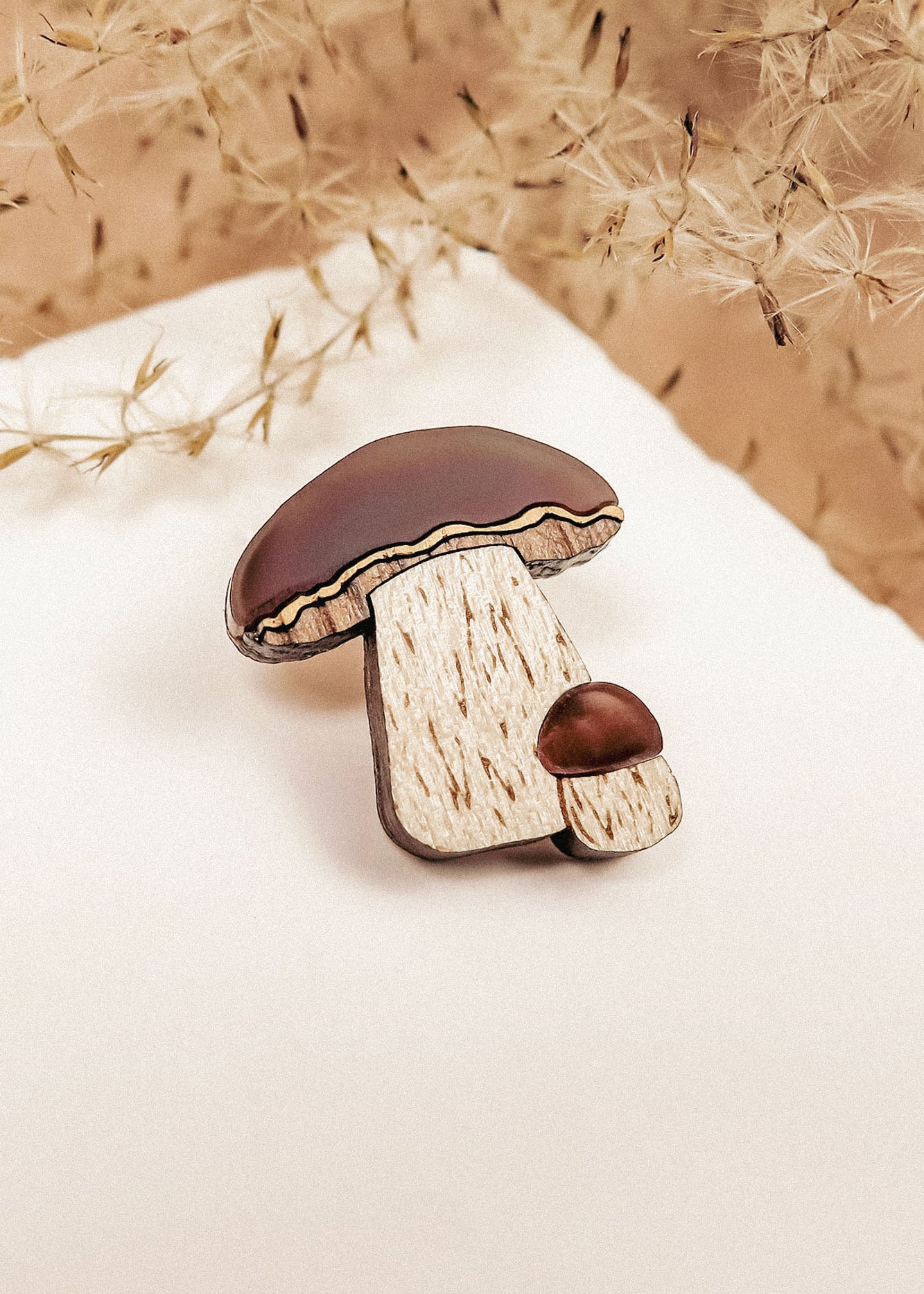 A wooden brooch featuring a nature-inspired design of two brown-capped mushrooms, one large and one small, placed on white paper with dried beige plants in the background. Showcases handcrafted quality and resembles a King Bolete Mushroom Pin.