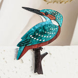 A colorful handcrafted Kingfisher Brooch featuring turquoise, brown, and white details perches on a textured white surface, with blurred beige foliage in the background.