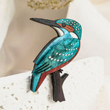 A colorful Kingfisher Brooch, handcrafted from sustainable materials, features blue, teal, brown, and white details as it perches on a sleek black branch against a light, textured background with blurred greenery.