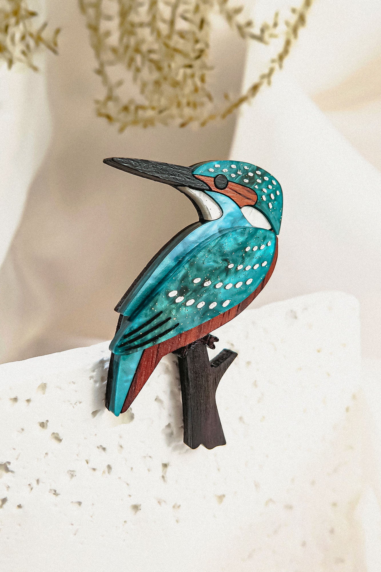 A colorful Kingfisher Brooch, handcrafted from sustainable materials, features blue, teal, brown, and white details as it perches on a sleek black branch against a light, textured background with blurred greenery.