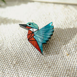 A kingfisher pin brooch shaped like a bird with geometric patterns in teal, red, black, and brown, crafted from recycled wood and placed on textured beige fabric with soft natural light.