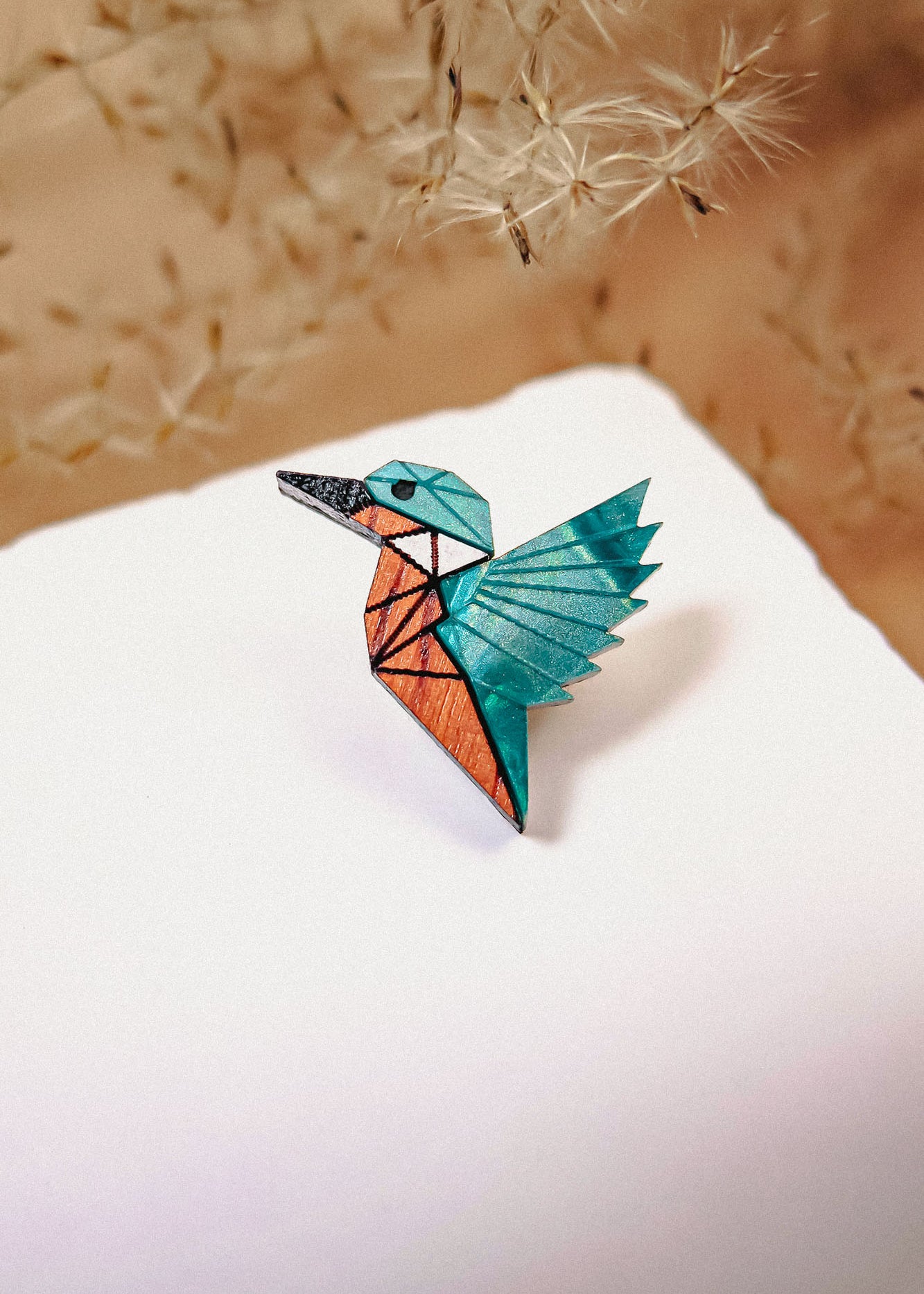 A colorful origami bird crafted from eco-friendly materials with blue, teal, and orange sections sits on a white surface, delicate dried grass or dandelion seeds softly blurred in the background.