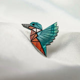 A colorful, geometric brooch shaped like a hummingbird rests on a white, softly textured surface. Crafted from eco-friendly materials, the brooch features teal, orange, and brown sections with black outlines in a stained-glass effect.