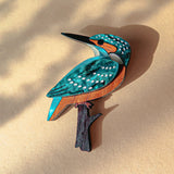 A colorful, handcrafted Kingfisher Brooch made from sustainable materials rests on a beige surface with soft shadows. The wooden brooch features striking blue, teal, white, and brown details.