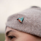 A close-up of a person wearing a knit beige hat decorated with a colorful kingfisher pin in shades of teal, blue, and orange. This eco-friendly accessory is perfect for bird lovers. The person's eyebrow and eye are partially visible.