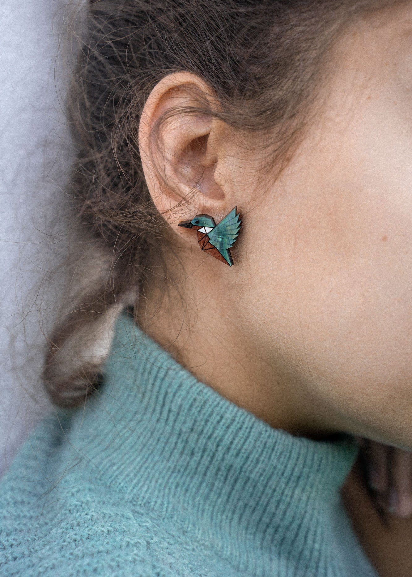Close-up of a person wearing a turquoise sweater and eco-friendly Kingfisher Stud Earrings, shaped like a hummingbird, with the focus on their ear and earring. The background is softly blurred.