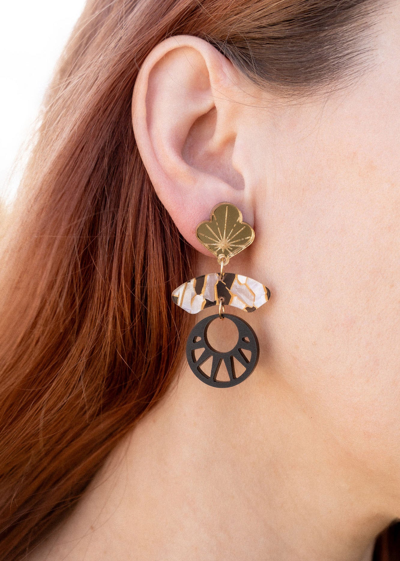 Close-up of a person with long auburn hair wearing handcrafted Kintsugi Earrings featuring a gold leaf, a clear crystal, and a black circular charm with a cut-out sunburst design inspired by Japanese art.