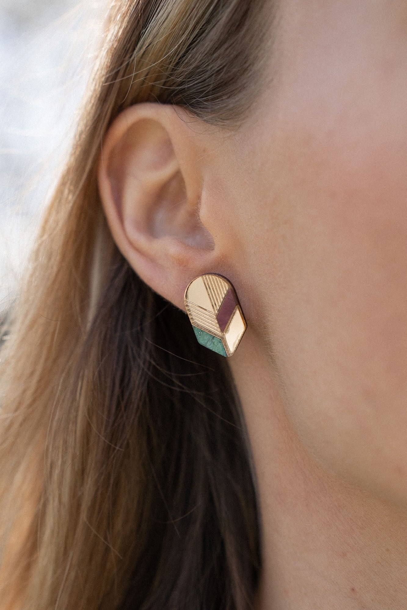 Close-up of a womans ear wearing a geometric, gold-toned stud earring with pastel accents. This piece of minimalist jewelry stands out against the softly blurred background, highlighting its unique handmade design and part of her hair and face.