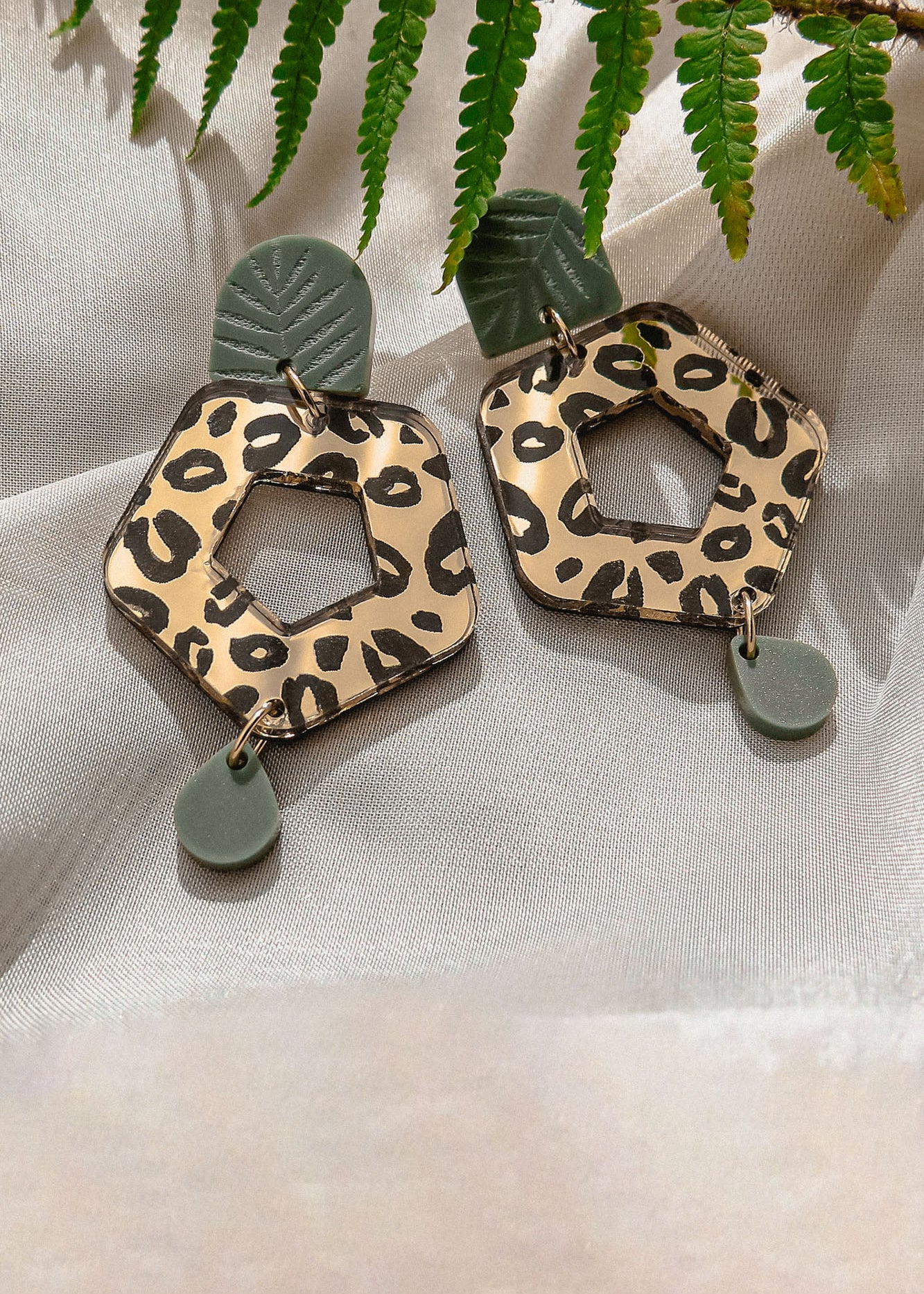 A pair of Leopard Earrings featuring timeless patterns—leopard print hexagons with green leaf-like and circular accents—crafted from premium materials, displayed on light fabric with a fern leaf above.