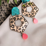 A pair of Leopard Earrings featuring leopard print hexagons, turquoise and black leaf-patterned tops, and dangling pink teardrops—crafted from premium materials, displayed on white fabric with a green fern leaf nearby.