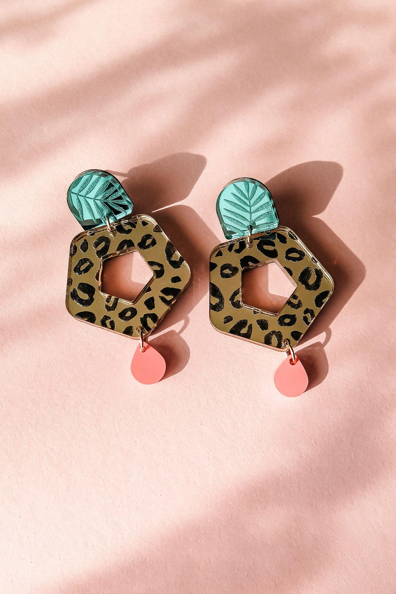 A pair of Leopard Earrings featuring green leaf-shaped tops, hexagonal leopard-print centers with cutouts, and small pink teardrop dangles—displayed on a light pink background for standout style with timeless patterns.