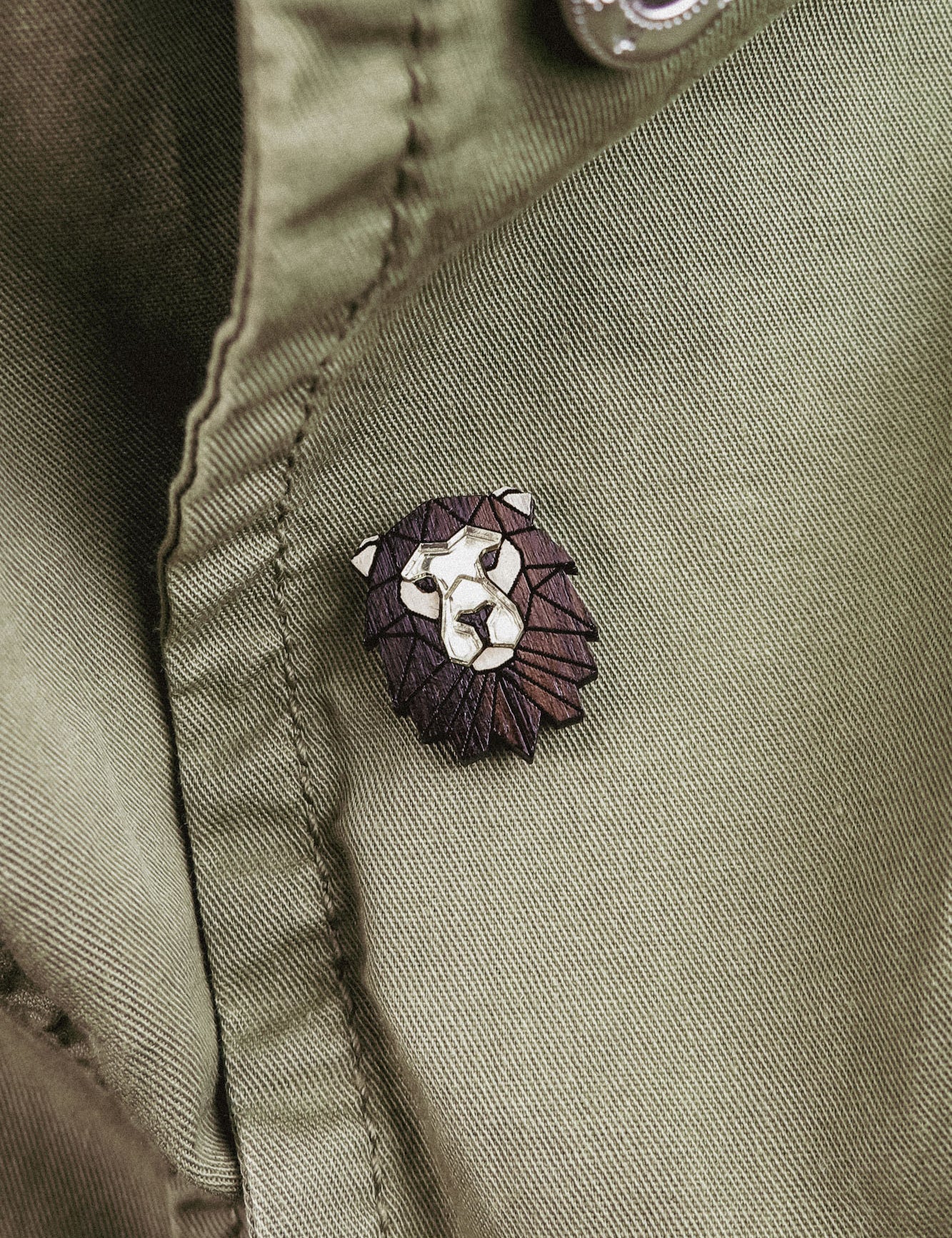 A close-up of an olive green jacket with a handcrafted geometric accessory—a Lion Pin—attached near a button.