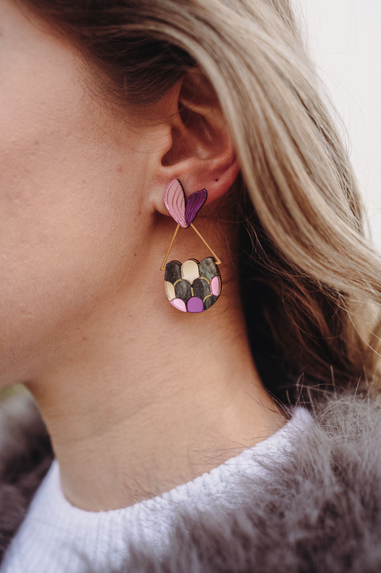 Close-up of a woman's ear wearing playful Mermaid Tail Earrings with a pink and purple heart-shaped stud and a semi-circular dangle in green, pink, and purple—a perfect accessory for mermaid lovers. The woman has blonde hair and wears a white top.