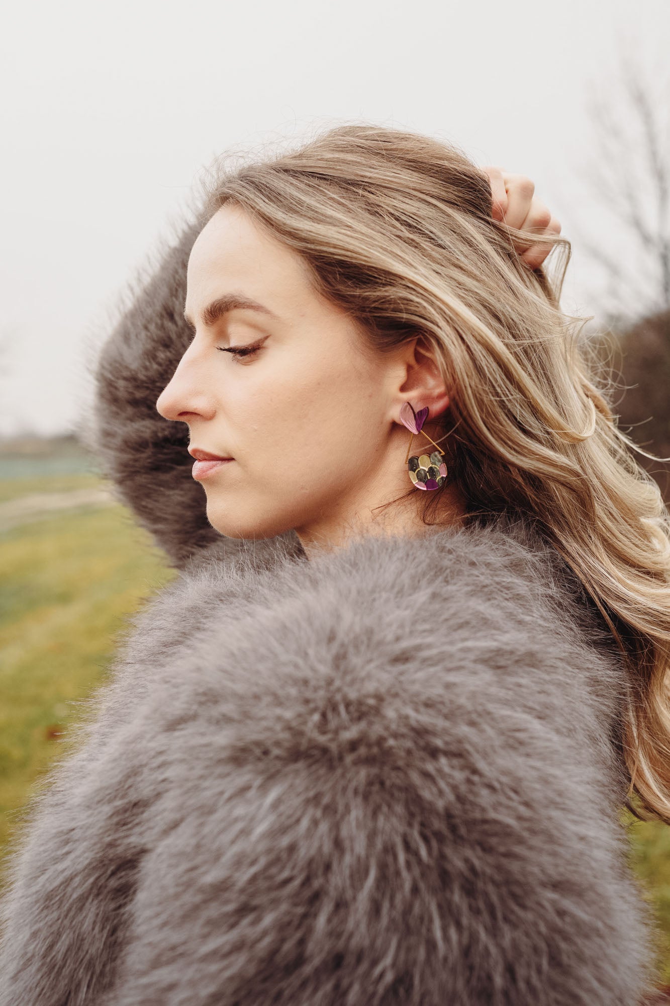 A woman with wavy blonde hair, wearing a fluffy gray coat and playful Mermaid Tail Earrings, stands outdoors with her eyes closed and one hand in her hair. The background is grassy with blurred trees—perfect for mermaid lovers.