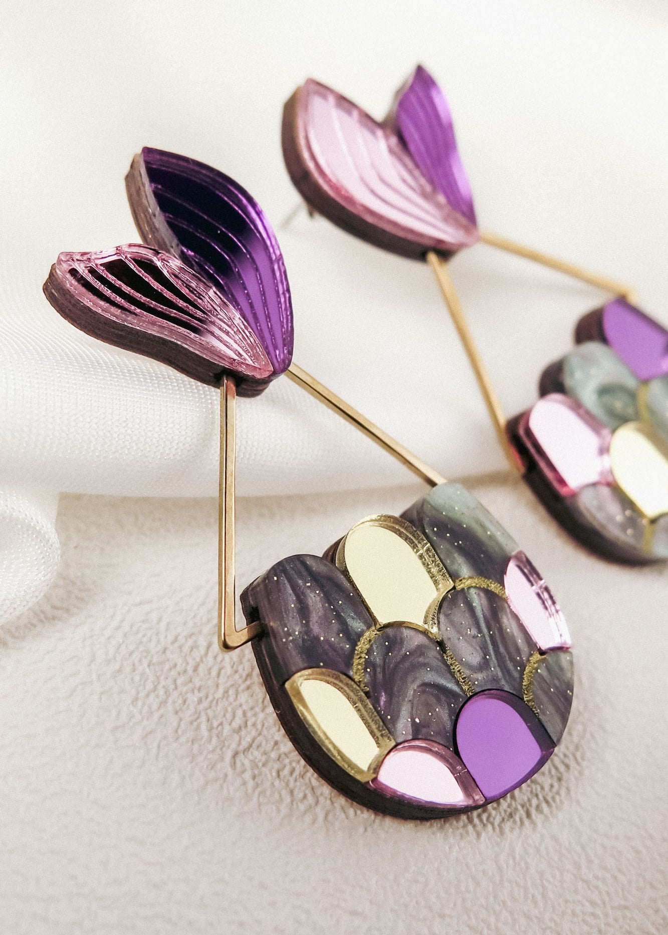 Colorful Mermaid Tail Earrings with a fan-shaped purple top and a semi-circular base featuring metallic and pastel scales in gold, pink, and gray, perfect for mermaid lovers seeking whimsical elegance. Displayed on a soft, textured white surface.