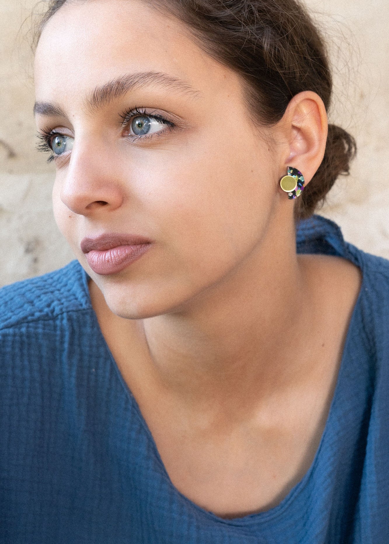 A young woman with light eyes and brown hair pulled back, wearing a textured blue top and lightweight Minimalist Geometric Stud Earrings, looks thoughtfully to the side against a beige, textured wall.