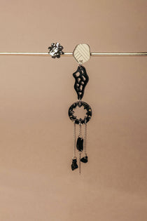 A single handcrafted earring featuring black and silver geometric shapes and dangling chains hangs from a horizontal metallic rod against a beige background—perfect for those who love lightweight earrings with an abstract flair.