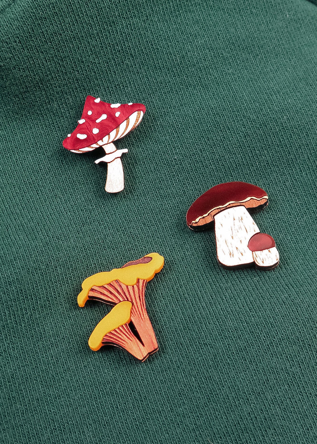 Three colorful, handcrafted enamel pins shaped like different mushrooms—including a Morel Mushroom Pin—are attached to a green fabric surface. These vibrant pins are made using eco-friendly materials for a sustainable touch.