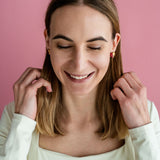 A woman with shoulder-length brown hair and a white top smiles with her eyes closed against a pink background, touching her hair with both hands, while wearing delicate handcrafted earrings.