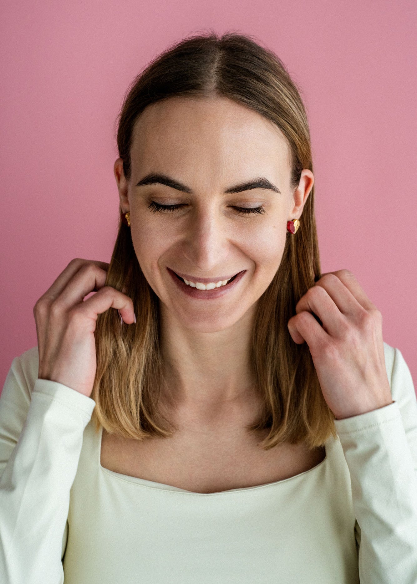 A woman with shoulder-length brown hair and a white top smiles with her eyes closed against a pink background, touching her hair with both hands, while wearing delicate handcrafted earrings.