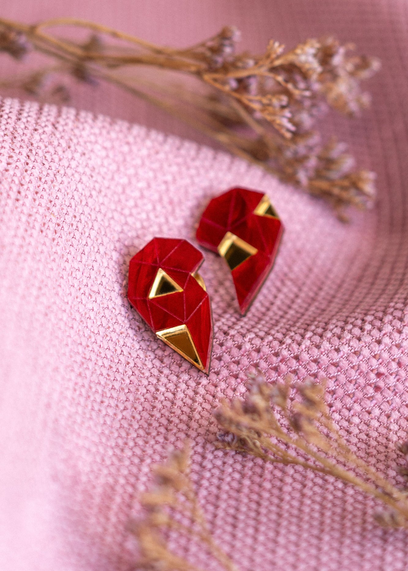 A pair of handcrafted geometric red and gold earrings shaped like hearts rest on textured pink fabric, with small dried flowers placed nearby—a perfect romantic gift.