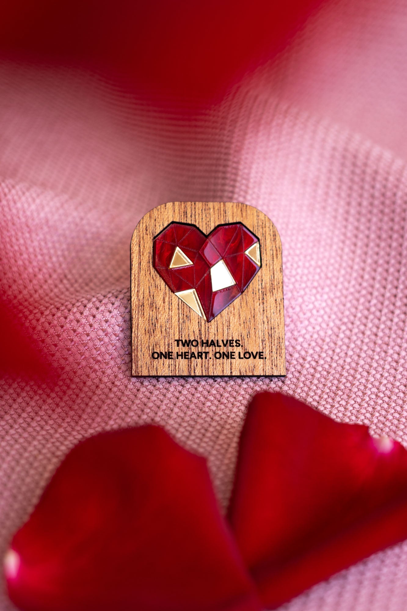 A handcrafted wooden pin with a red geometric heart and the words TWO HALVES. ONE HEART. ONE LOVE. rests on pink textured fabric, surrounded by rose petals—a romantic gift from the One Love Halves Pins collection.