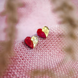 A pair of handcrafted red and gold geometric heart-shaped earrings rest on a textured pink fabric, with blurry plant stems in the foreground—perfect romantic jewelry for any occasion.