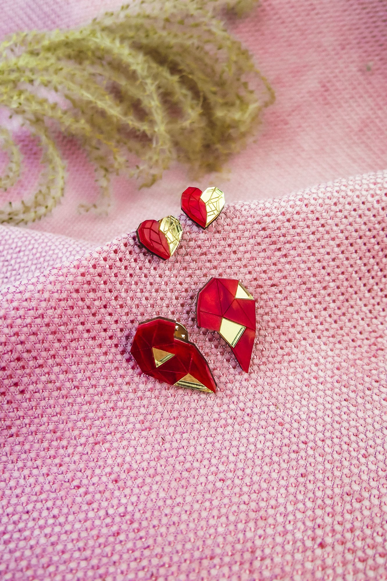 Red and gold geometric heart studs and pins, perfect as romantic jewelry, are displayed on textured pink fabric, with decorative plant stems in the background.
