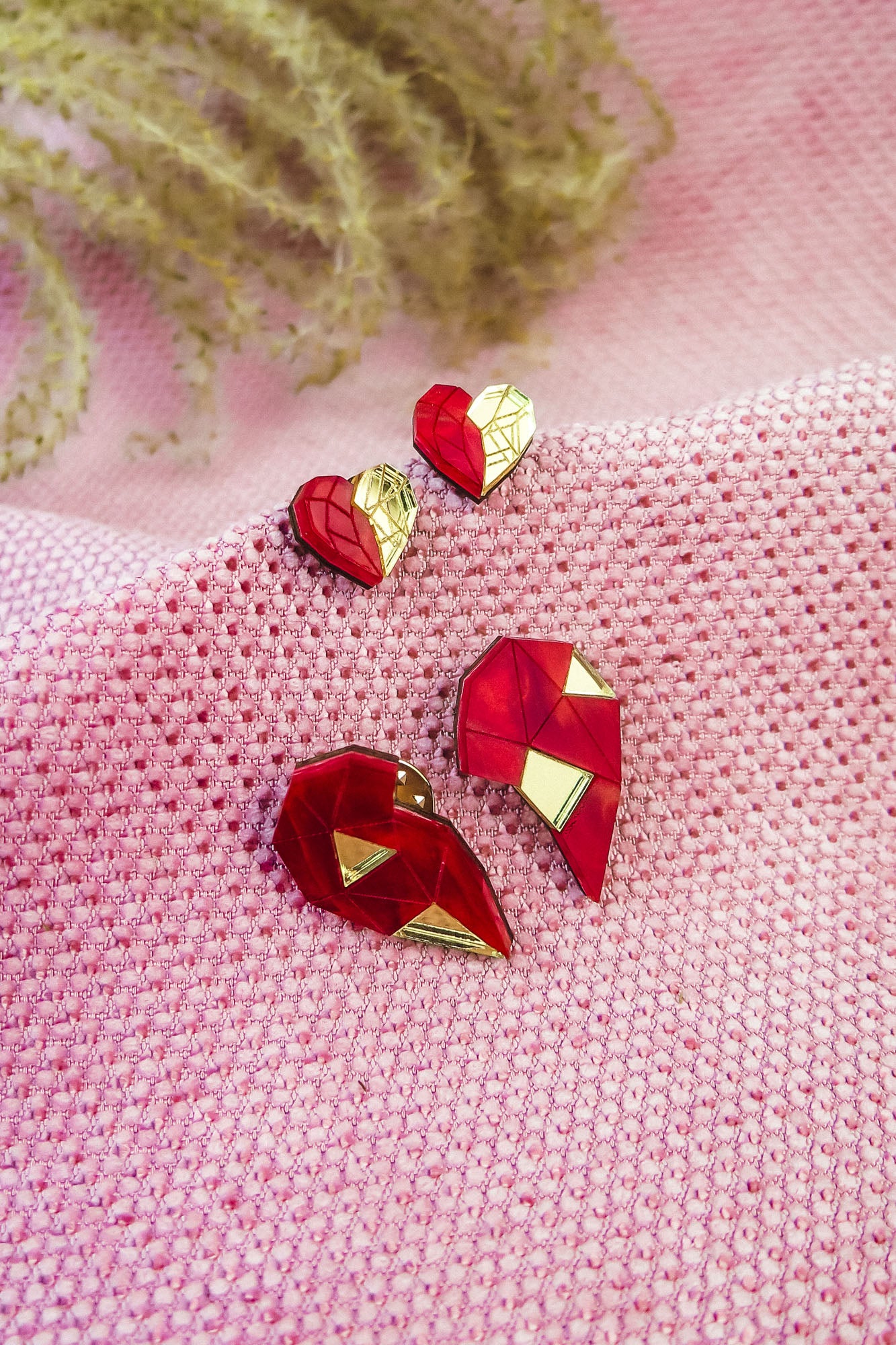 Red and gold geometric heart studs and pins, perfect as romantic jewelry, are displayed on textured pink fabric, with decorative plant stems in the background.