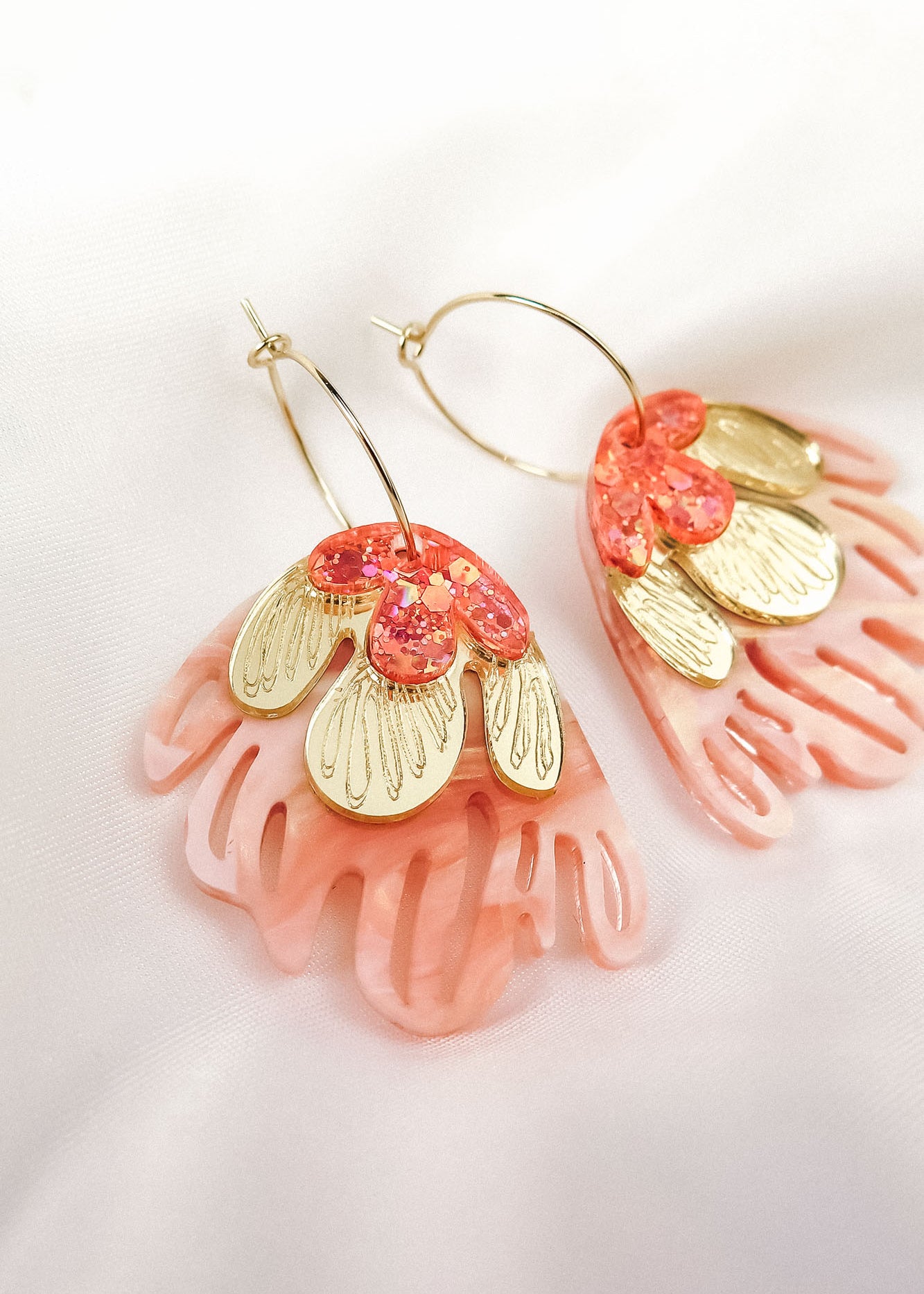 A pair of Organic Floral Hoops with pink, coral, and gold petal-like acrylic flowers rest on a soft white fabric background. These gold hoops feature intricate cut-out designs and shimmering accents for a delicate, nature-inspired look.