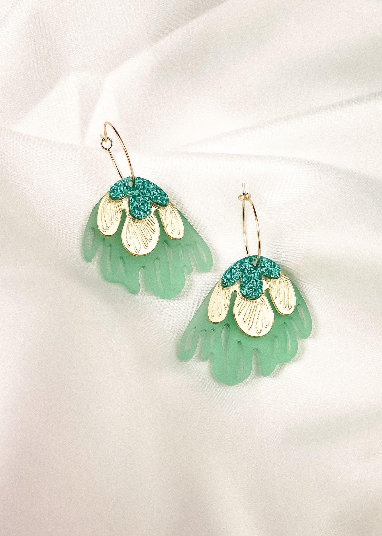 A pair of handcrafted dangling earrings with gold hooks, featuring layered designs in mint green, cream, and teal glitter, resembling abstract floral shapes for a touch of tropical elegance, displayed on a white fabric background.