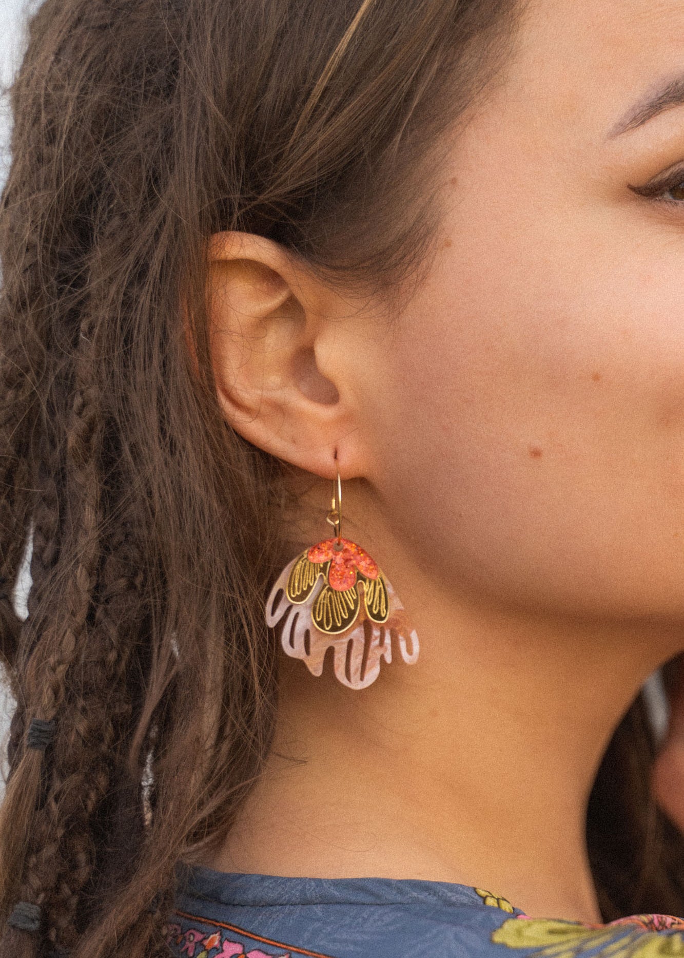 A close-up of a woman’s face with brown skin and dreadlocks, wearing vibrant Organic Floral Hoops adorned with pink, gold, and red acrylic flowers shaped like leaves or petals. The background is softly out of focus.