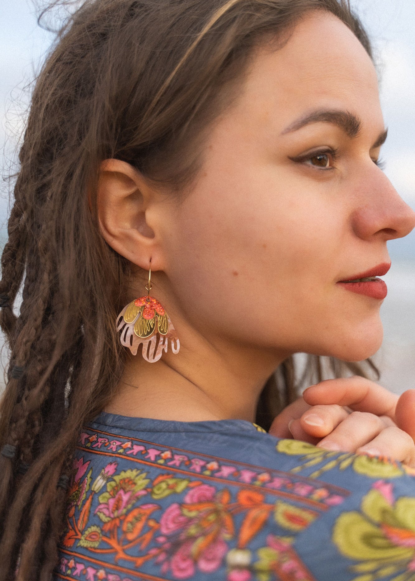 A woman with brown dreadlocks and winged eyeliner looks to the side, wearing a colorful floral-patterned garment and striking Organic Floral Hoops with pink and gold accents. She has a slight smile, her hands gently clasped.