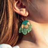 Close-up of a person’s ear wearing a large, colorful handcrafted earring shaped like a stylized flower with gold, turquoise, and light green petals. The person has light skin and long, light brown hair.