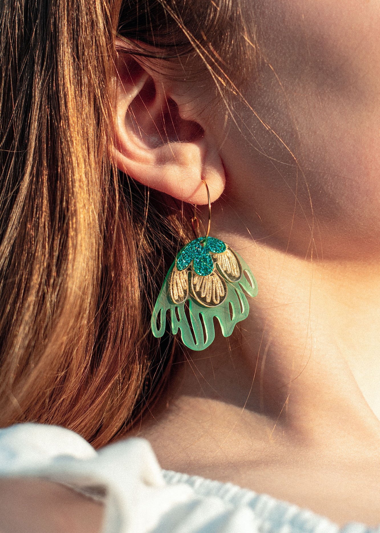 Close-up of a person’s ear wearing a large, colorful handcrafted earring shaped like a stylized flower with gold, turquoise, and light green petals. The person has light skin and long, light brown hair.