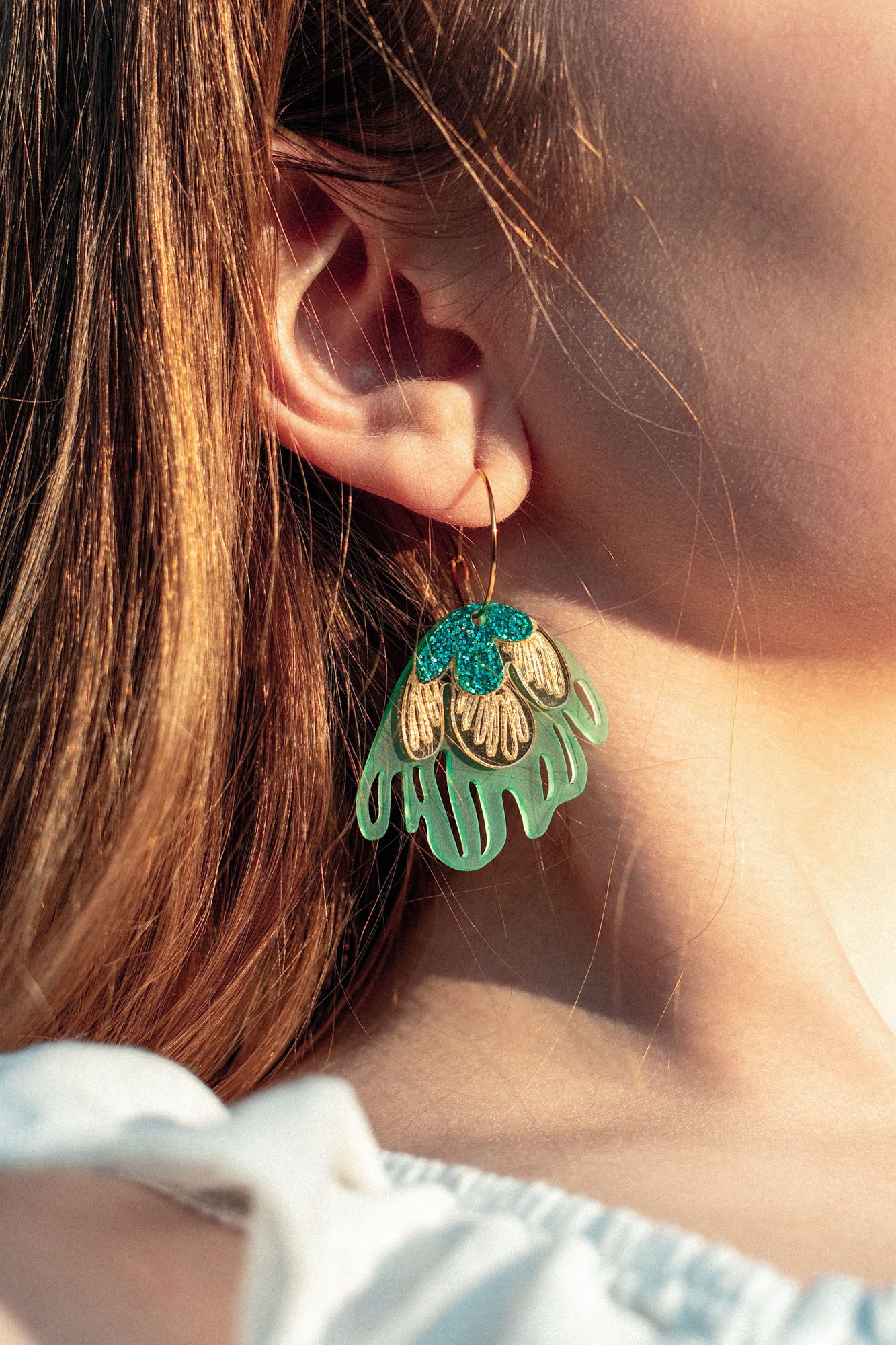Close-up of a person’s ear wearing a large, colorful handcrafted earring shaped like a stylized flower with gold, turquoise, and light green petals. The person has light skin and long, light brown hair.
