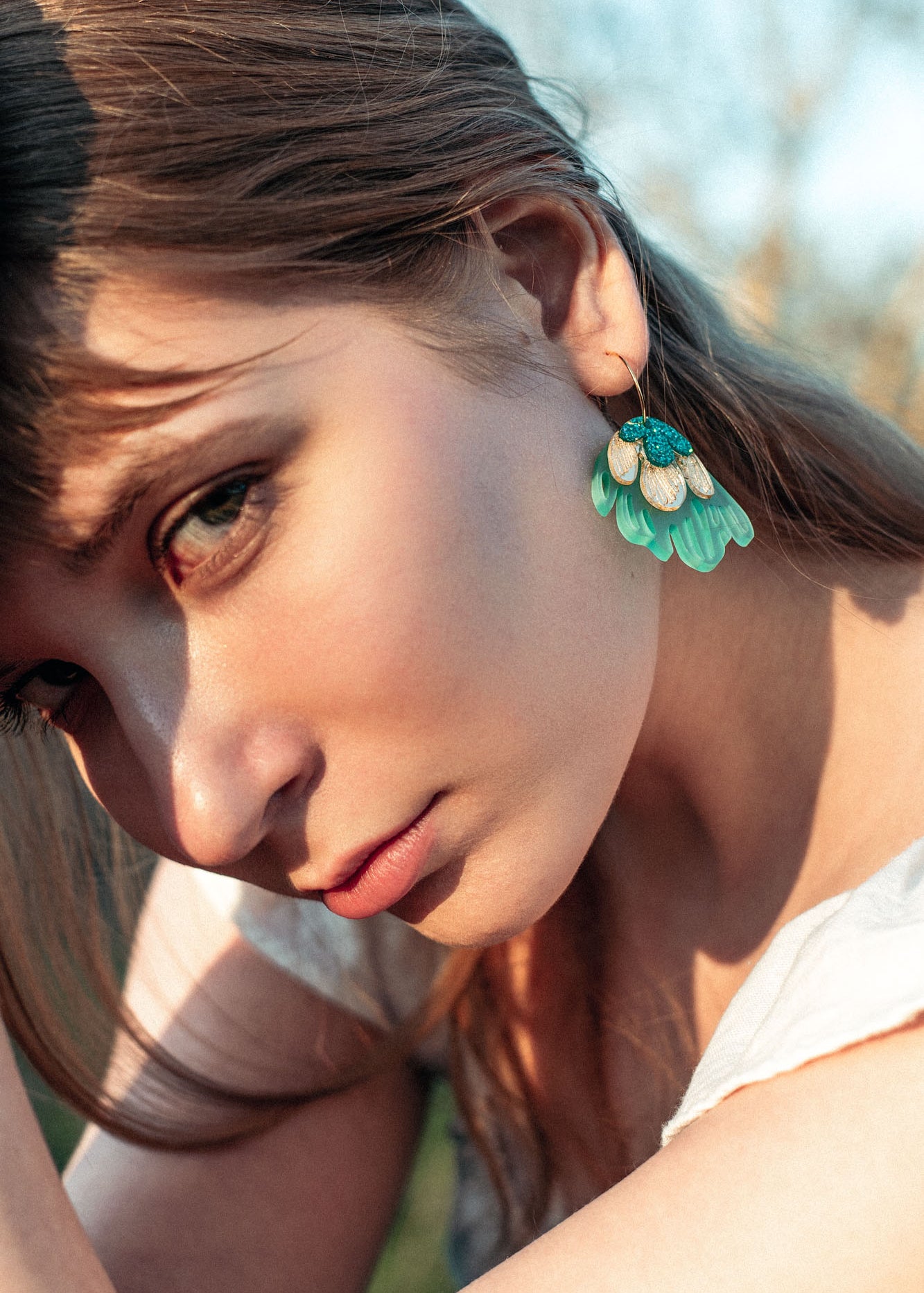 A young woman with long brown hair gazes intently at the camera, wearing a white top and handcrafted earrings—Organic Floral Hoops with turquoise and gold leaf accents. Natural outdoor lighting highlights her face.