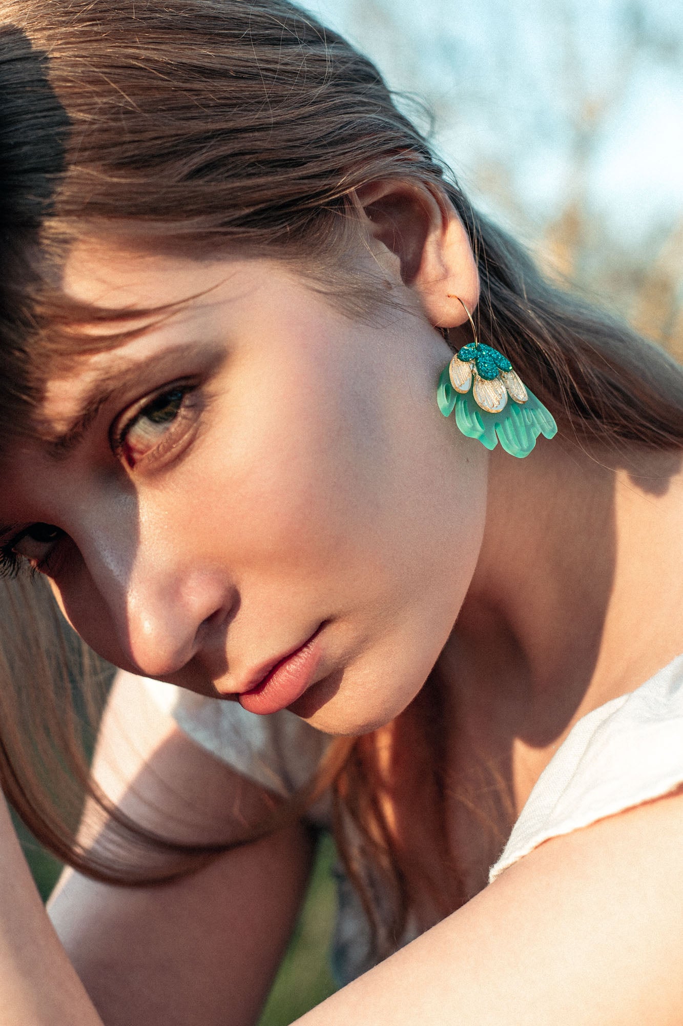 A young woman with long brown hair gazes intently at the camera, wearing a white top and handcrafted earrings—Organic Floral Hoops with turquoise and gold leaf accents. Natural outdoor lighting highlights her face.