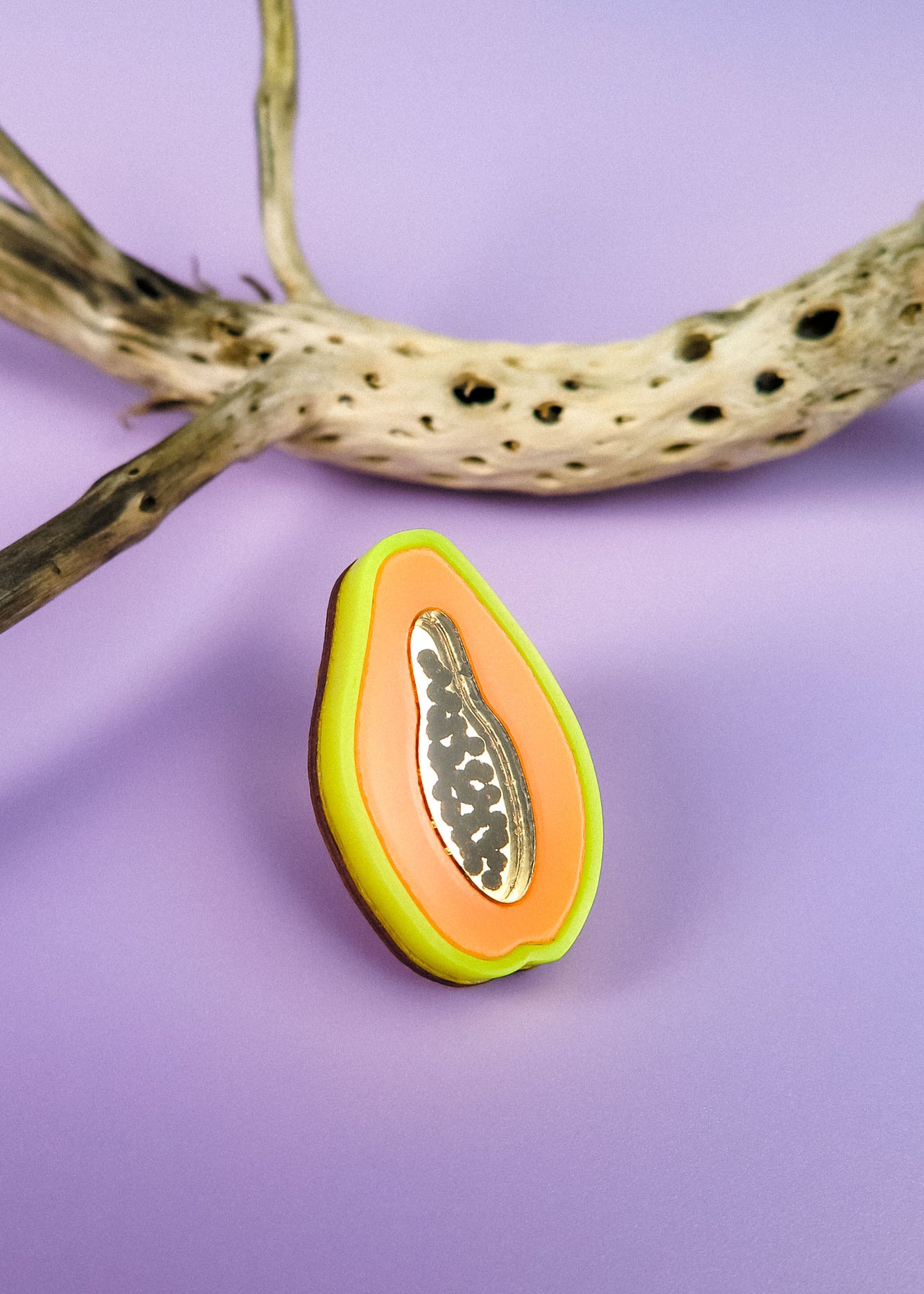 A handcrafted, colorful papaya-shaped cookie with icing details sits on a purple surface next to a dried, spotted branch, reminiscent of a neon Papaya Pin.
