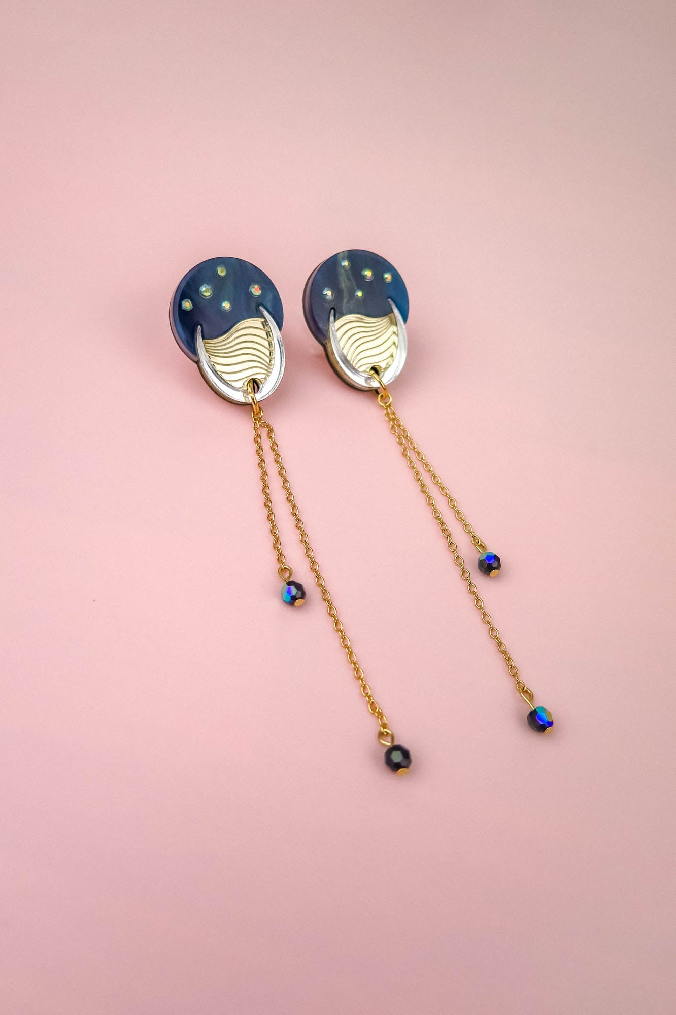 A pair of earrings with blue circular tops reminiscent of the night sky, gold accents, and long gold chains with small blue beads, displayed on a pink background.