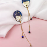 Two gold and blue earrings with dangling chains and beads rest on a white fabric, set against a pink background, evoking a hint of festival magic.