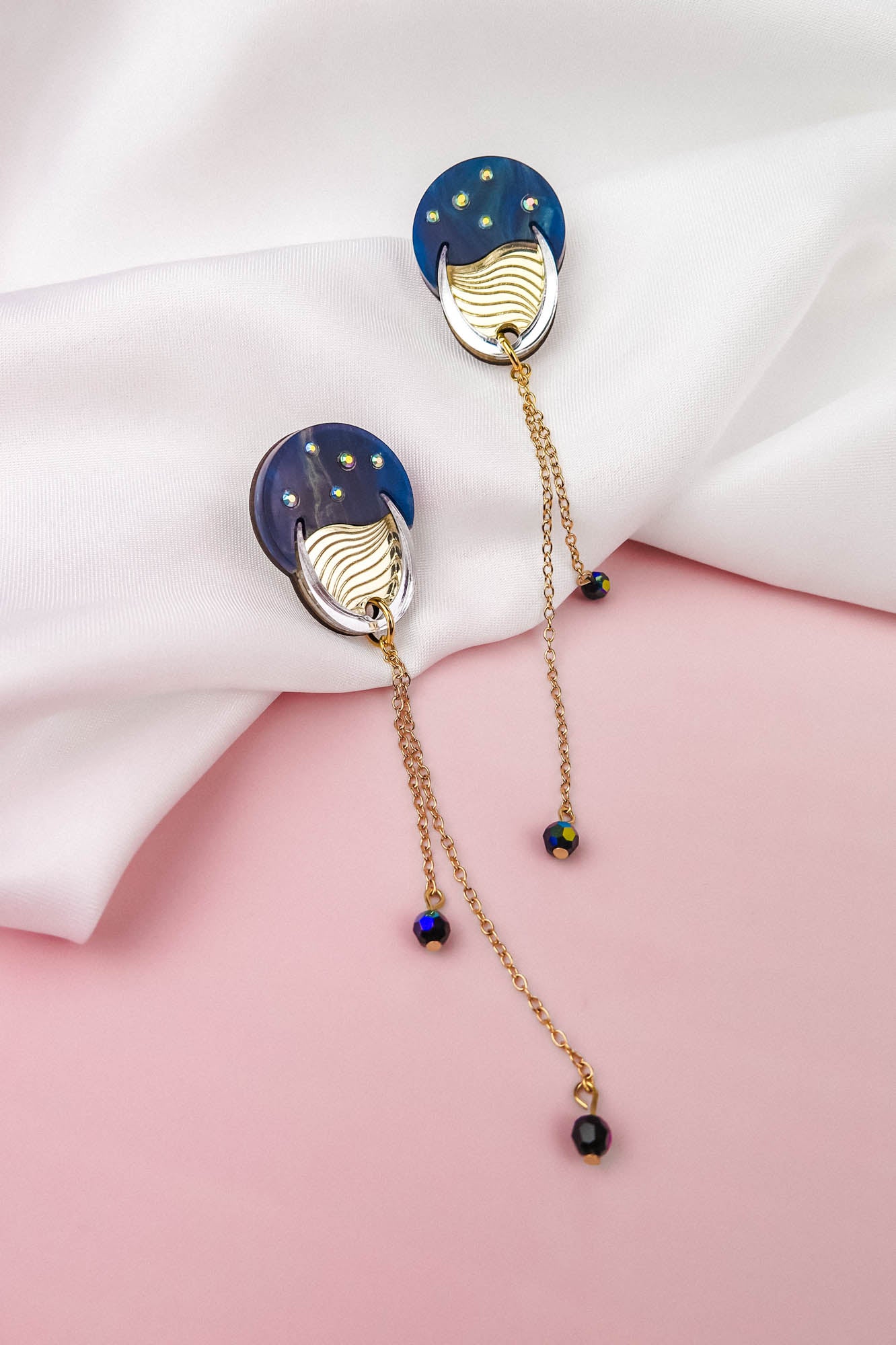 Two gold and blue earrings with dangling chains and beads rest on a white fabric, set against a pink background, evoking a hint of festival magic.