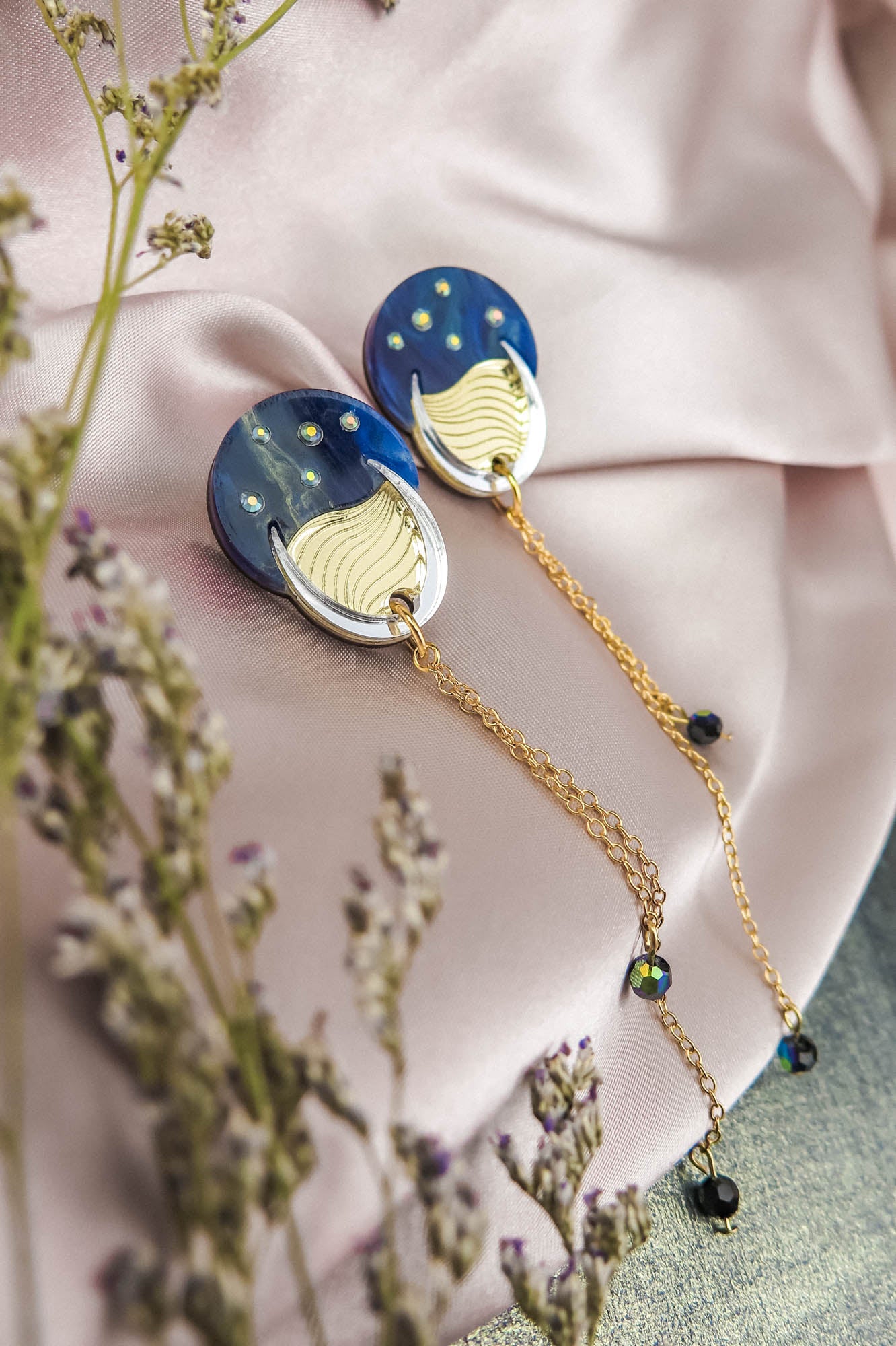 Two round, blue and gold earrings reminiscent of the night sky, with dangling gold chains, are placed on a light pink satin fabric. Small flowers with purple buds frame the foreground, adding to the festival magic of the scene.